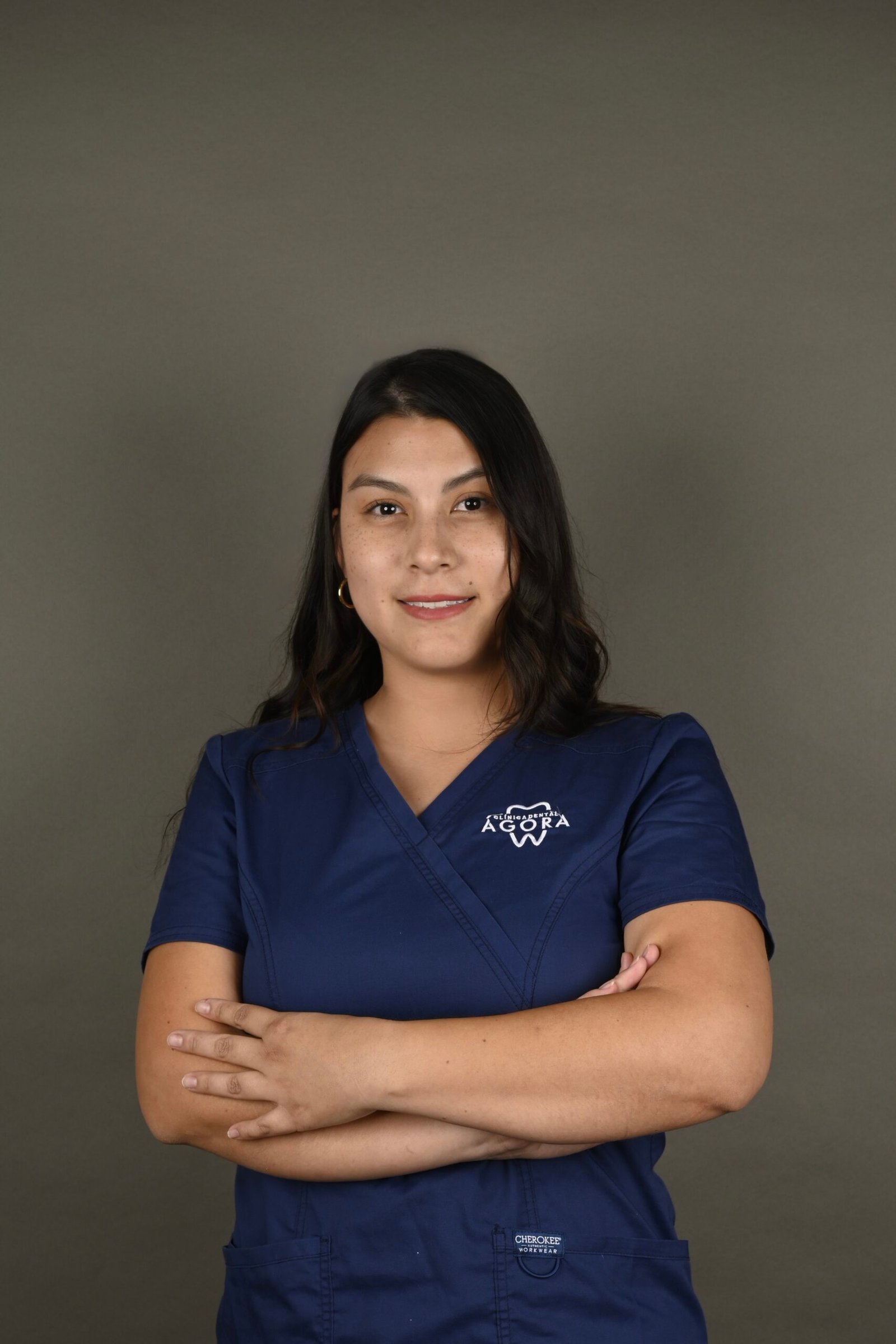 Retrato profesional de Érika Montoya Cardona, higienista dental en Clínica Dental Ágora, con uniforme clínico y los brazos cruzados frente a un fondo gris